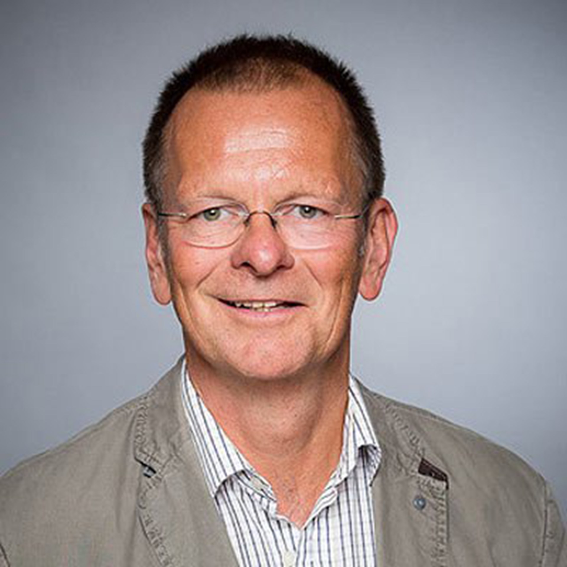 Lächelnder Mann mit kurzen, blonden Haaren und Brille, der ein helles, gestreiftes Hemd und eine hellgraue Jacke trägt